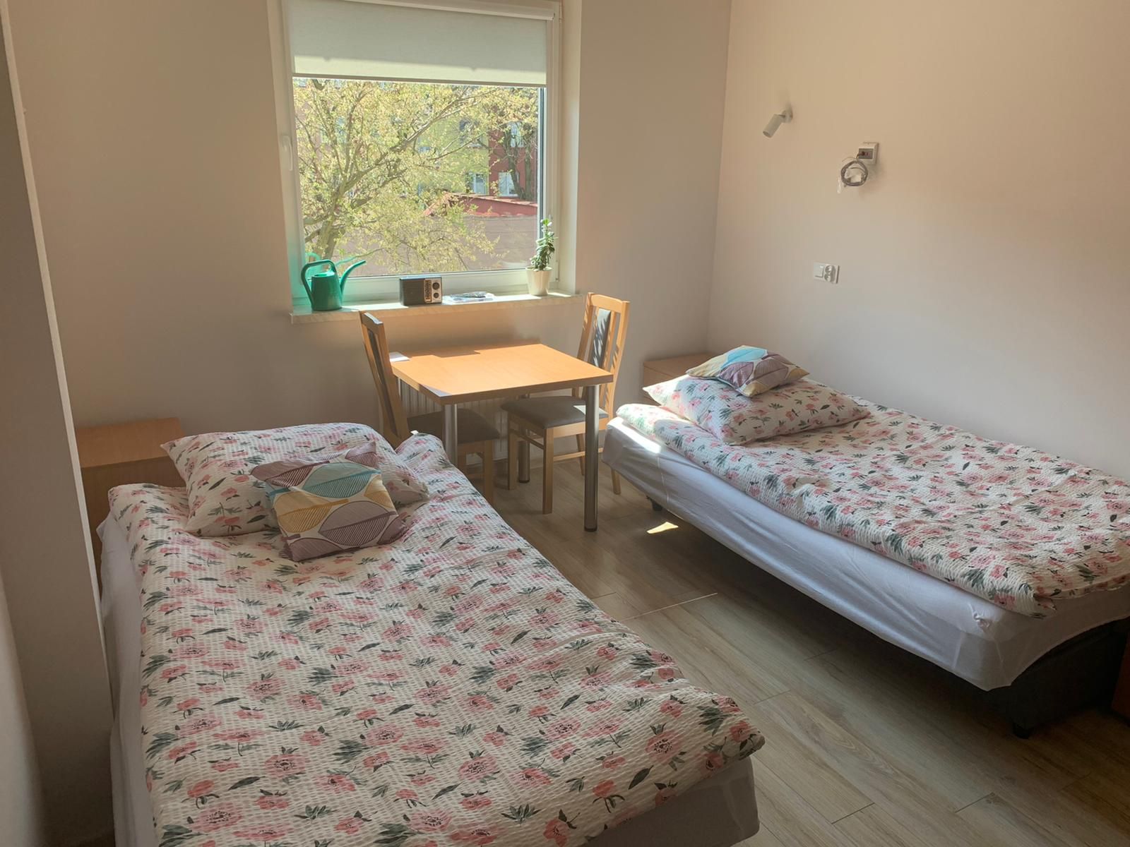 Jasny pokój dwuosobowy w DPS „Ostoja”: dwa łóżka, stolik z krzesłami przy oknie